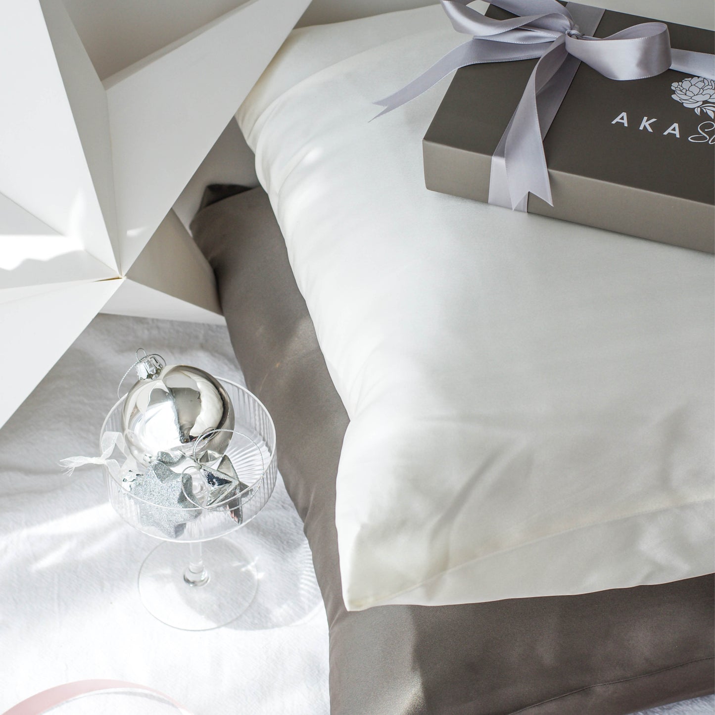 AKA Sleep Mullberry Silk pillowcases with Night Slate on the bottom and Moon white on top. Pillows are on top of a white linen sheet. Top left corner is a white cardboard star. Bottom left is a champagne coupe filled with silver baubles. Top right corner is a charcoal AKA Sleep box wrapped with a light charcoal silver ribbon.