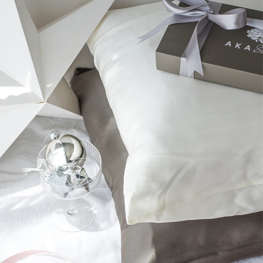 AKA Sleep Mullberry Silk pillowcases with Night Slate on the bottom and Moon white on top. Pillows are on top of a white linen sheet. Top left corner is a white cardboard star. Bottom left is a champagne coupe filled with silver baubles. Top right corner is a charcoal AKA Sleep box wrapped with a light charcoal silver ribbon.